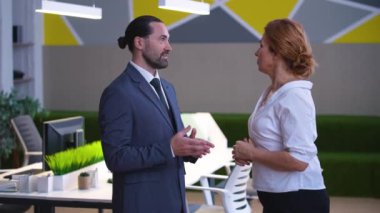 A stylish office, a man in a suit and a woman in a shirt, talk, discuss work, solve issues. Colleagues, a businessman stand sideways to the camera