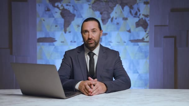 穿着西服打领带的时髦男子坐在桌旁 现代笔记本电脑 向摄像机讲述新闻 背景是一张世界地图 — 图库视频影像