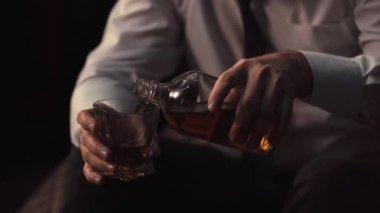 Takım elbiseli ve kravatlı bir adam evde bir otel odasında oturur ve alkol döker. İş adamı viski içer, yorucu bir günün ardından viski içer, yorgundur, kamera hareket eder.