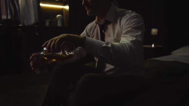 一个穿衬衫的男人坐在旅馆的床上 手里拿着一瓶酒 威士忌 威士忌 威士忌 白兰地 商人喝酒 下班后疲倦 — 图库视频影像