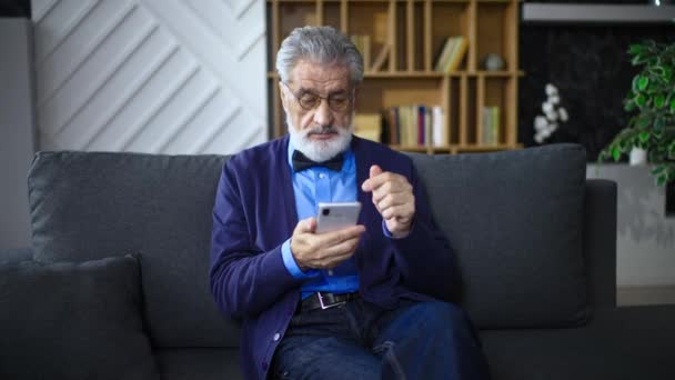 一位穿着衬衫 手拿着电话打着领结的成年男子正坐在沙发上 随随便便放松了下来 老人笑着 滚动着电话 在家里看新闻 — 图库视频影像