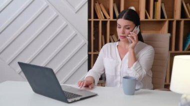 Genç bir kadın dizüstü bilgisayarın önünde oturmuş telefonda konuşuyor. Memnuniyetle gülümsüyor ve hafta sonu için planlardan memnun. Ofis kamerasında.