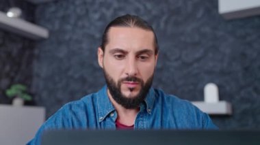 Evdeki ofiste dizüstü bilgisayarla çalışan konsantre olmuş genç bir adamın portresi. Kot gömlekli genç sakallı bir iş adamı modern bir dizüstü bilgisayar üzerinde yoğunlaşıyor.