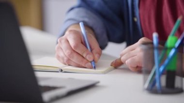 Masada serbest çalışan bir öğretmen dizüstü bilgisayarda yazı yazıyor. İnternetteki Webinar kursunu yakından izle. Erkeklerin elleri bir deftere modern bir dizüstü bilgisayar tüketicisi ürünleri yazıyor.