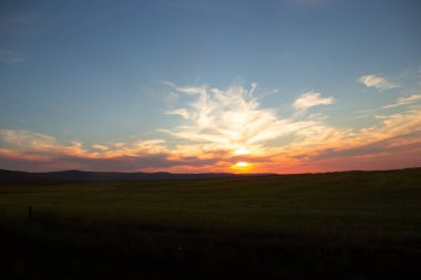 Feather clouds above the prairies and hills in the orange sunset time. Sun is behind clouds.