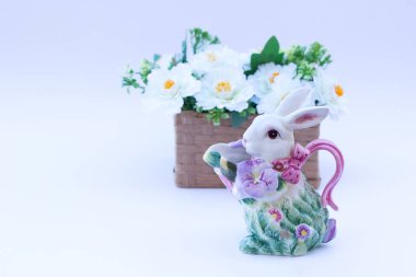 Paskalya dekoru. Renkli tavşancık şekilli seramik fincan tabağı. Arka planda beyaz çiçekler olan hasır sepetin yanında..