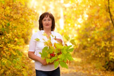 Beyaz gömlekli emekli kadın, sonbahar parkında sarı yapraklarla yol kenarında dikiliyor..