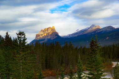 Bulutlu bir sonbahar gününde, nehrin yukarısındaki derin ladin ormanlarının arasında, sabah ışığında yüksek bir dağ. Castle Dağı, Banff Ulusal Parkı, Kanada.