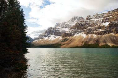 Soğuk bir sonbahar gününde bulutlu gökyüzü ile gölün üzerindeki karla kaplı dağlar turkuaz sularla kaplıdır. Bow Gölü, Banff Ulusal Parkı, Kanada.