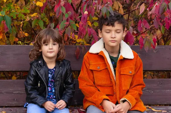Two kids, toddler girl and her older brother are sitting sad and bored on the bench in the fall park with bright foliage.