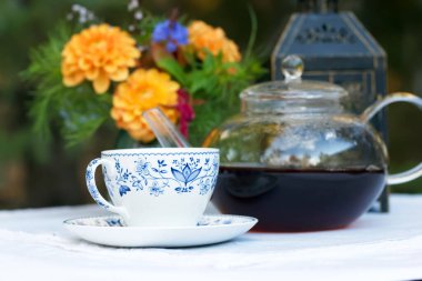 Mavi çiçekli beyaz bir fincan çay ve beyaz kumaşlı teras masasında şeffaf bir demlik, vazoda çiçekler ve arka planda bir fener..