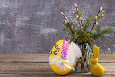 Easter composition with yellow bunny figurine and decorated egg near the vase with pussy willow branches and evergreen plants (labrador tea, bear berry, fir) on the wooden brown table.