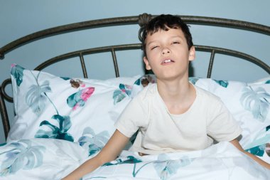 Uykulu, mutsuz bir çocuk, yorgun bir yüz ifadesiyle yatakta oturuyor. Metal yatak başlığı ve yapraklı beyaz ve yeşil yatak..