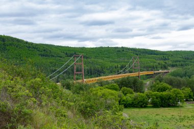 Yellow metal bridge over the river surrounded with green woodlands on hills in summer, blue cloudy sky. Dunvegan bridge crossing Peace River, Alberta, Canada. clipart