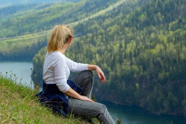 Genç kadın kanyon tepesinde oturuyor ve yazın nehir, dağ ve ormanın manzarasına hayran..