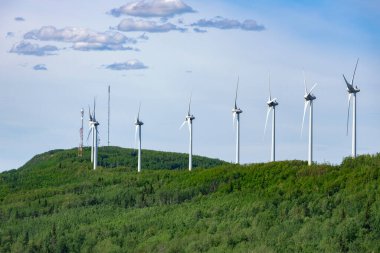 Rüzgâr türbinlerinin oluşturduğu sıra, yaz mevsiminde yeşil ağaçlar ve mavi gökyüzü arasında dağda sürdürülebilir enerji üretir..