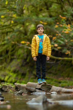 Gündelik giysiler içinde beyaz bir çocuk sonbaharda yağmur ormanlarının nemli deresinde doğanın tadını çıkarıyor. Kayalarda yeşil yosun ve ağaçlarda sarı yapraklar. Vancouver Adası, British Columbia, Kanada.