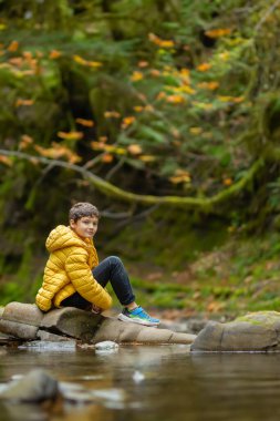 Gündelik giysiler içinde beyaz bir çocuk sonbaharda yağmur ormanlarının nemli deresinde doğanın tadını çıkarıyor. Kayalarda yeşil yosun ve ağaçlarda sarı yapraklar. Vancouver Adası, British Columbia, Kanada.