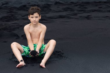 Renkli mayo giymiş genç bir çocuk, siyah kumdaki suyun kenarında oturuyor ve elinde bir avuç dolusu volkanik kum tutuyor. Black sand Beach, Big Island, Hawaii, ABD.
