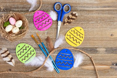 Renkli ahşap yumurtalar, boya fırçaları, makas, şeker, benekli yumurta ve tüylerle dolu kahverengi ahşap masa üzerinde canlı Paskalya sanatı kompozisyonu.