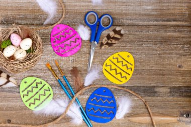 Renkli ahşap yumurtalar, boya fırçaları, makas, şeker, benekli yumurta ve tüylerle dolu kahverengi ahşap masa üzerinde canlı Paskalya sanatı kompozisyonu.