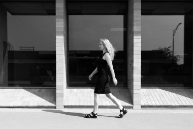 Siyah-beyaz bir fotoğraf. Siyah elbiseli genç bir kadın. Şehir caddesinde beton ve cam bina geçerek yürüyor..