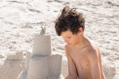 Beyaz tenli genç çocuk okyanus kıyısında beyaz bir kumsalda oturuyor ve yaz tatillerinde kumdan kale inşa ediyor..