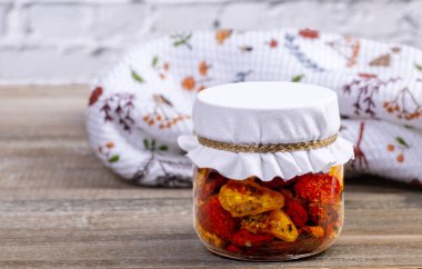 Kurumuş kırmızı ve sarı kiraz domatesli zeytinyağı ve bitkilerle dolu cam bir kavanoz. Kavanoz, arka planda mutfak havlusu olan kahverengi ahşap masa..