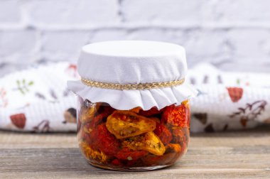 Kurumuş kırmızı ve sarı kiraz domatesli zeytinyağı ve bitkilerle dolu cam bir kavanoz. Kavanoz, arka planda mutfak havlusu olan kahverengi ahşap masa..