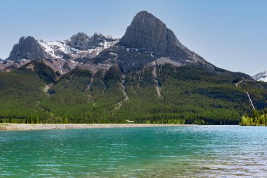 Berrak turkuaz suları olan bir gölün dağlarında güzel bir manzara, yeşil ağaçlarla kaplı dağ tepesinde kayalar, güneşli bir yaz günü. Rundle Forebay, Banff Ulusal Parkı, Kanada.