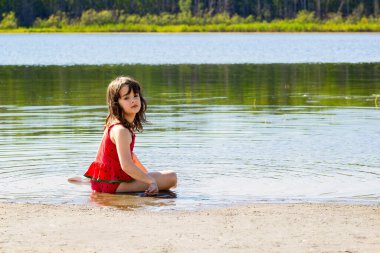 Küçük kız, sıcak yaz gününde, kırsal gölün sakin sularında kumsalda oturuyor..