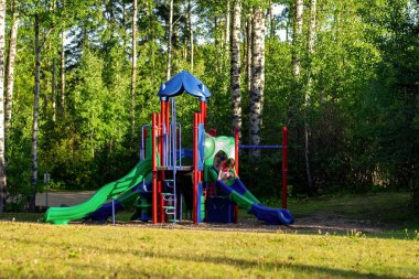 Yaz mevsiminde yeşil parkta kaydırak, merdiven, merdiven ve salıncakla parlak renkli bir oyun alanı. Küçük kız kaydırağından kayıyor.