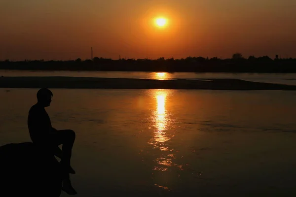 一个人坐在河边岩石上看着日落的轮廓 — 图库照片