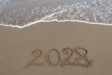 Yeni yıl 2028 deniz dalgası arka planında kumsalda yazılmış..
