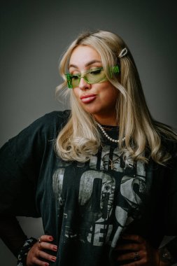 Yeşil gözlüklü ve koyu renk elbiseli sarışın bir kız stüdyoda koyu bir arka planda fotoğraf çekiliyor.