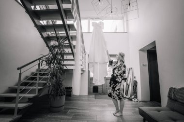 Narin bir gelinlik evin koridorundaki güzel bir korkuluğa asılır. Gelin sabah eteği içinde elbisenin yanında duruyor. Güzel ışık. Düğün fotoğrafı. Yüksek kalite