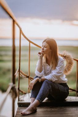 Bir kadın gün batımında kot pantolon ve beyaz bir gömlekle gölün kıyısında oturuyor.