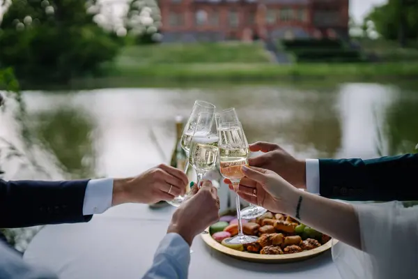 Four people clinking champagne glasses close-up at the table, castle and lake in the background People are nibbling on each other outside