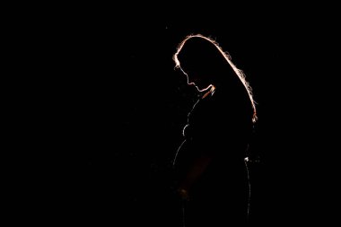 Düşük tuşlu hafif portrede, güzel hamile bir kadının göbeğinin silüeti siyah bir arkaplanda göze çarpar, çarpıcı ve anımsatıcı bir görüntü yaratır.