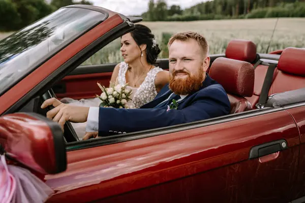 In a vibrant red convertible, a newlywed couple sits, radiating joy and happiness in a delightful portrait of their special day