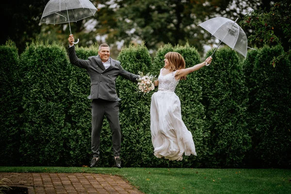 The happy couple jumps joyfully while holding a wedding umbrella, celebrating their special day with exuberance and love.