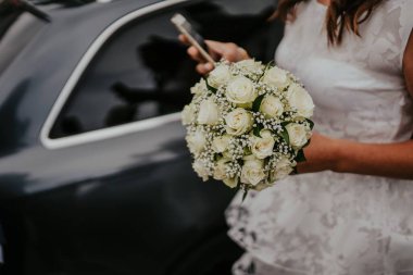 Bir buket beyaz çiçeğin yakın çekimi. Dantelli beyaz elbiseli gelin elinde bir buket tutuyor. Diğer elin akıllı telefonu var.