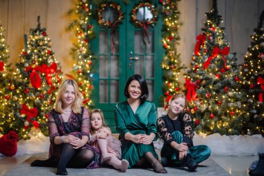 Üç kızı olan bir anne, Noel süslemeleri içinde oturmuş gülüyor, yeşil bir kapı ve bir sürü Noel ağacı yanında.