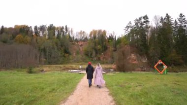 Biri siyah ceketli, diğeri pembe ceketli iki kişi, kumtaşı bir uçurum ve sonbahar ormanı olan çimenli bir bölgedeki ahşap köprüye doğru yürüyorlar.