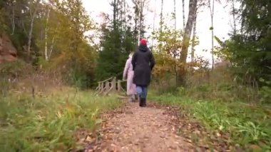 Biri kırmızı bereli, biri siyah ceketli, diğeri pembe ceketli iki kişi düşmüş yapraklarla kaplı bir orman yolunda yürüyorlar..