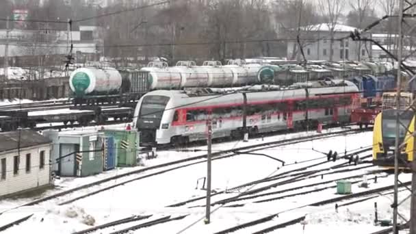 冬の間の鉄道ヤード 近代的な旅客列車は前景で見ることができ 部分的に頭上線に取り付けられています 雪でほこった線路に静止しているようだ — ストック動画
