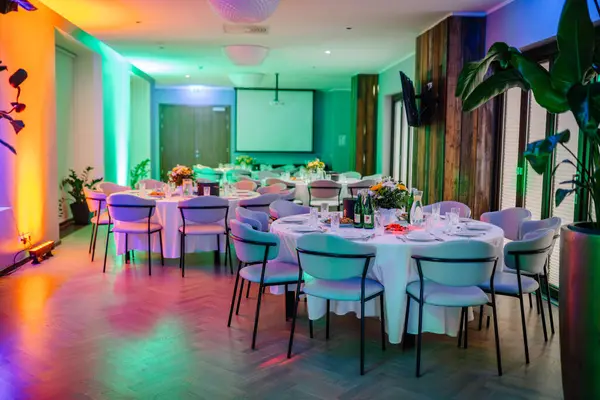 elegantly set event hall with round dining tables draped in white cloths, surrounded by modern blue chairs. The room is bathed in ambient multicolored lighting with a large screen on one wall