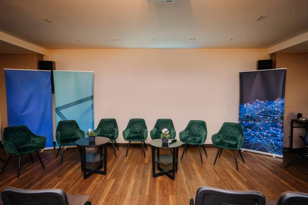 modern meeting room setup with a semi-circle of green velvet chairs around small round tables, each adorned with a plant, and abstract blue graphic roll-up banners in the background.