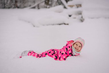 Karda oynarken gülümseyen neşeli bir şablona sahip canlı pembe bir kar giysisi içindeki çocuk. Kar taneleri yavaşça düşüyor..