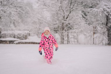Karda oynarken gülümseyen neşeli bir şablona sahip canlı pembe bir kar giysisi içindeki çocuk. Kar taneleri yavaşça düşüyor..